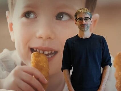 A man stands in front of a large picture of a boy eating a chicken nugget.  A man stands in front of a large picture of a boy eating a chicken nugget.