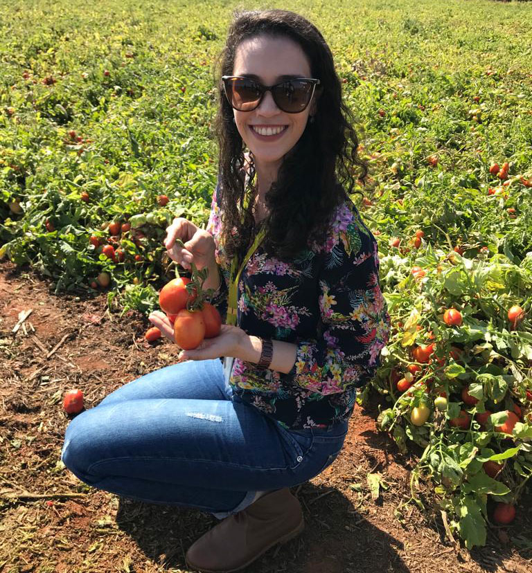 A woman smiles and holds up several tomatoes while kneeling down in front of rows of tomato plants.