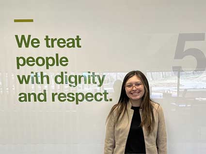 A young woman smiles in front of a wall with the message that says, ‘We treat people with dignity and respect.’