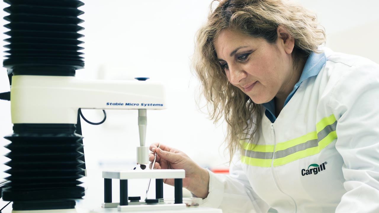 A woman in industrial garments observes a sample in a microscope. 