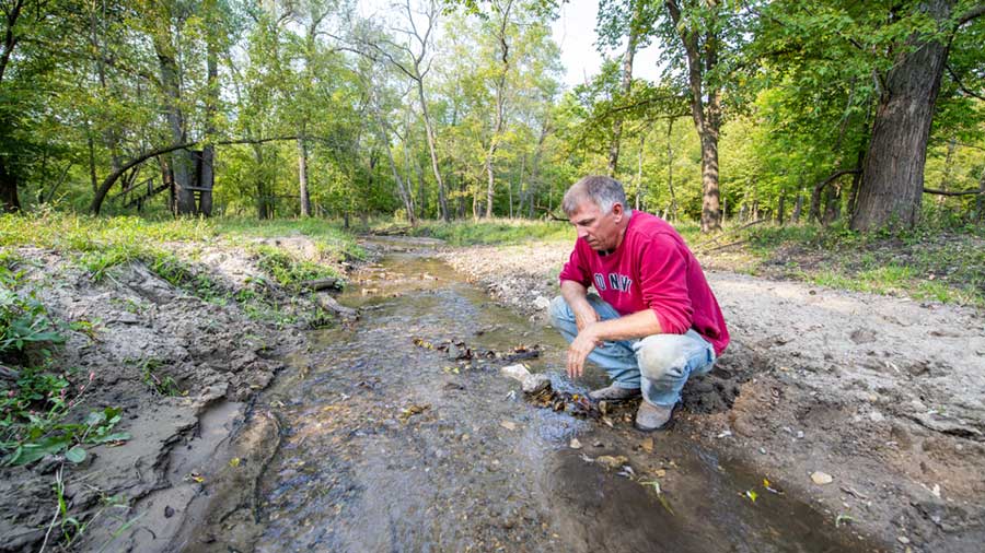 A farmer crouches down near a stream on his farmland. A farmer crouches down near a stream on his farmland.