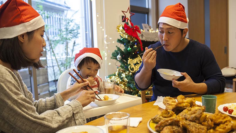 A woman, man and small child all wearing red holiday hats sit at a table with fried chicken and biscuits on it.  A woman, man and small child all wearing red holiday hats sit at a table with fried chicken and biscuits on it.