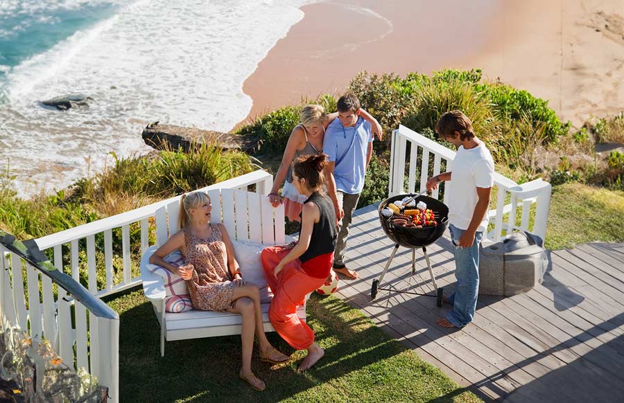 Five people stand around a bbq with a beach in the background. 