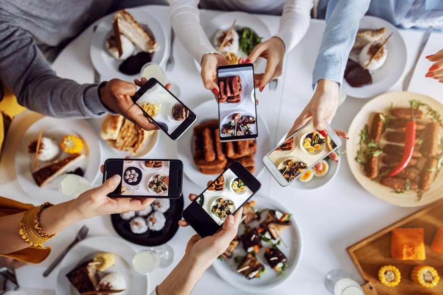 People snapping photos of food with smartphones around a table, showcasing plates filled with protein options like chicken and beef. People snapping photos of food with smartphones around a table, showcasing plates filled with protein options like chicken and beef.