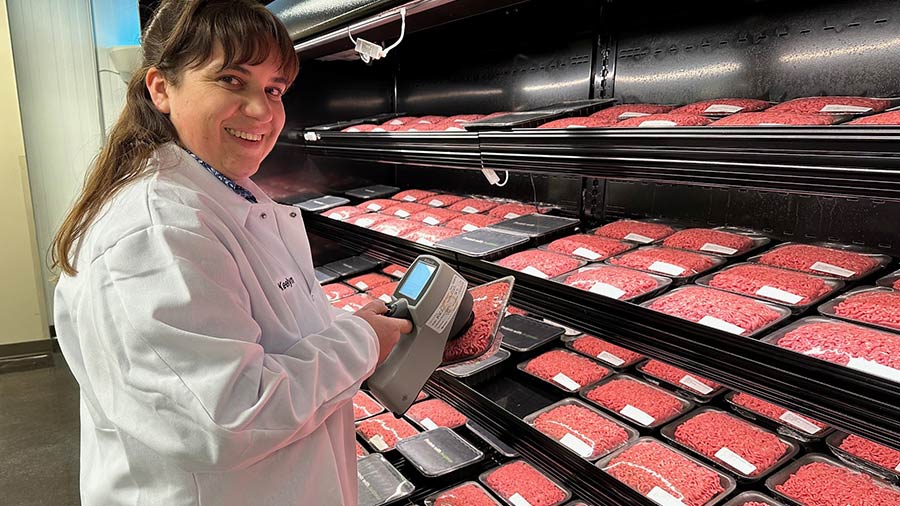 A Cargill employee in a white lab coat holds a scanner up to a package or red beef and smiles toward the camera. 