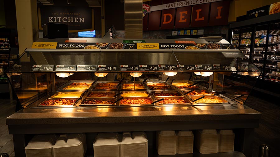 A grocery store hot food bar with trays of various cooked dishes under heat lamps, labeled with prices, and stacks of takeout containers below. A grocery store hot food bar with trays of various cooked dishes under heat lamps, labeled with prices, and stacks of takeout containers below.