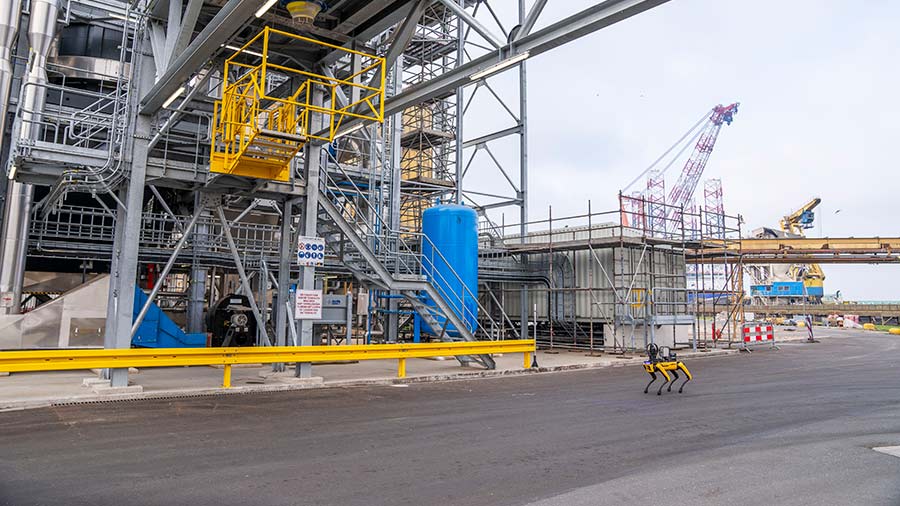 Spot the robotic dog walks on pavement outside a large operations facility. Spot the robotic dog walks on pavement outside a large operations facility.
