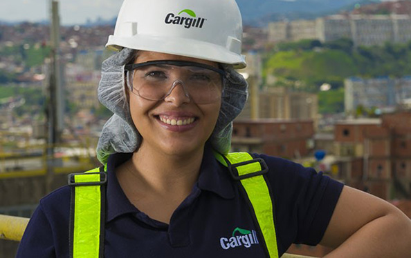 An employee in a white hard hat and yellow safety jacket stands over Spot the robotic dog as it sits on the ground in a plant.  An employee in a white hard hat and yellow safety jacket stands over Spot the robotic dog as it sits on the ground in a plant.