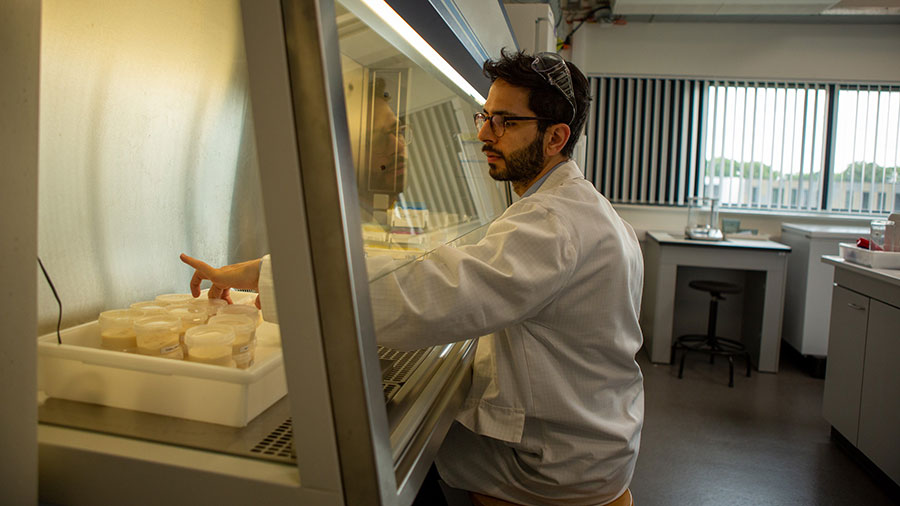 A man in a white lab coat and wearing glasses sits and moves a clear plastic cup filled with fermented liquid.  A man in a white lab coat and wearing glasses sits and moves a clear plastic cup filled with fermented liquid.