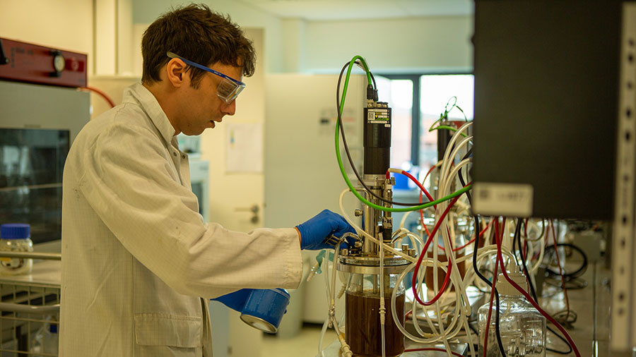 A man in a white lab coat wearing blue gloves works with a clear container of fermented liquid.  A man in a white lab coat wearing blue gloves works with a clear container of fermented liquid.