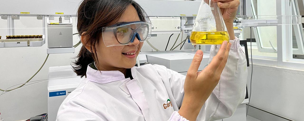 A woman wearing a white lab coat and safety glasses holds up a glass beaker with yellow liquid inside.  A woman wearing a white lab coat and safety glasses holds up a glass beaker with yellow liquid inside.