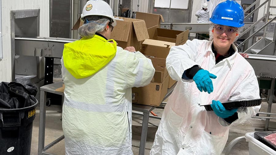 A woman wearing a white lab coat, blue hard hat and safety glasses smiles and looks toward the camera. 